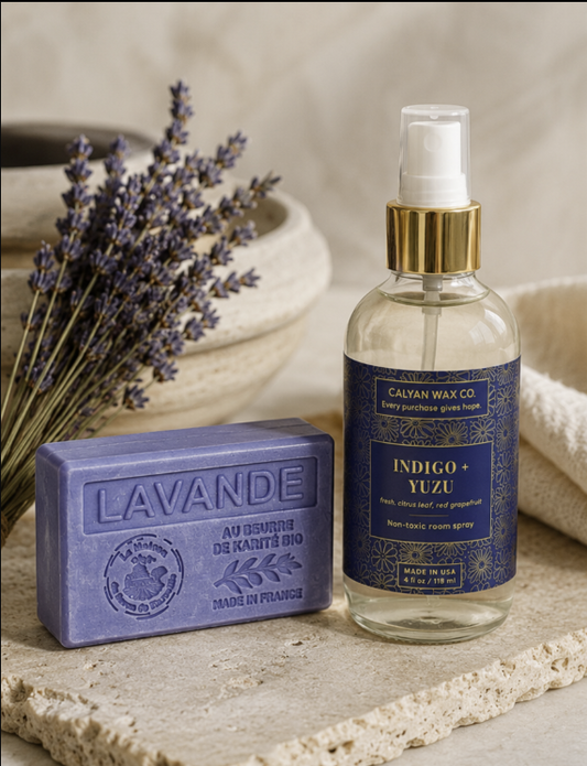 A bottle of Calyan Wax Co Indigo + Yuzu room spray and a lavender hand soap bar are styled on a stone surface with dried lavender and a neutral background.