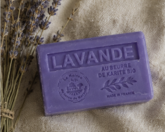 A lavender-colored rectangular bar of soap with embossed text reading 'LAVANDE' and 'AU BOURGEON DE KANATE BO', placed on a beige fabric surface with dried lavender sprigs at the side.